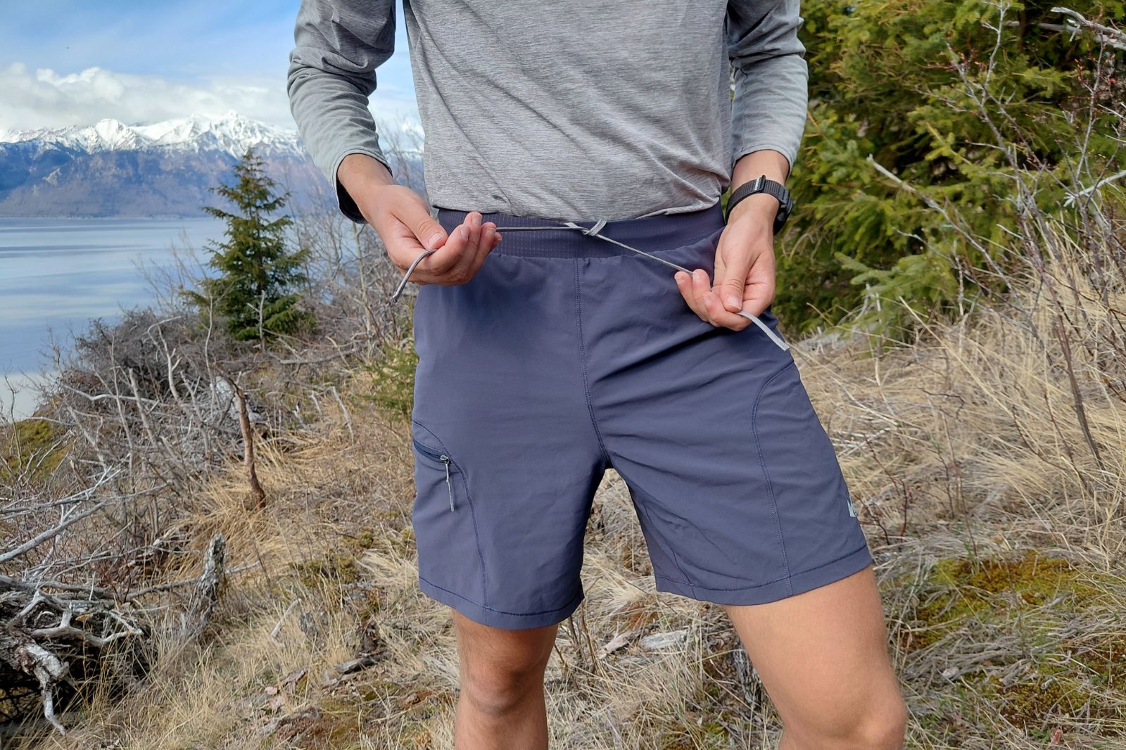 A hiker stands on a dirt trail on a hillside wearing a long-sleeve grey shirt and tying blue shorts.