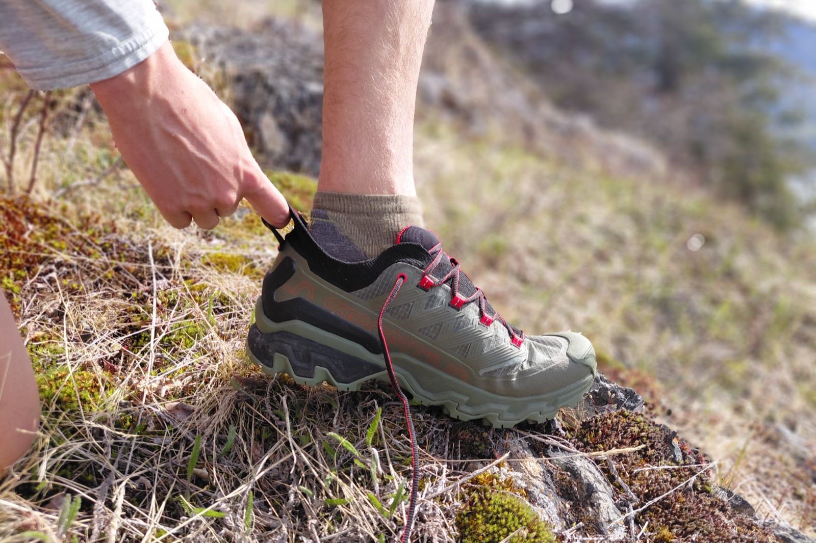 Close-up of a person adjusting an olive-green hiking shoe on rocky terrain.