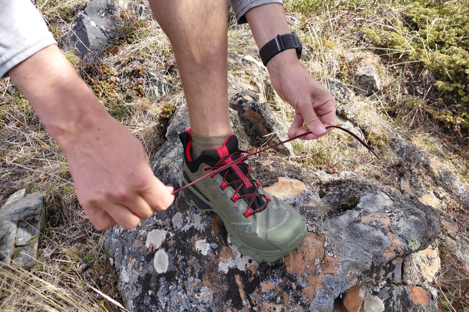 Person tying the laces of an olive green hiking shoe on rocky terrain.