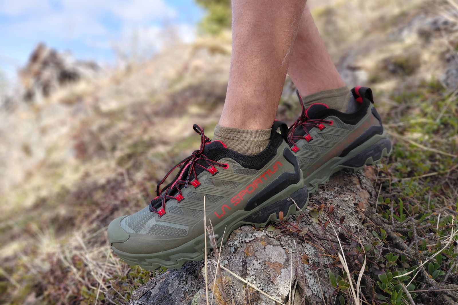 Person wearing olive green and black hiking shoes with red details on a rocky terrain.