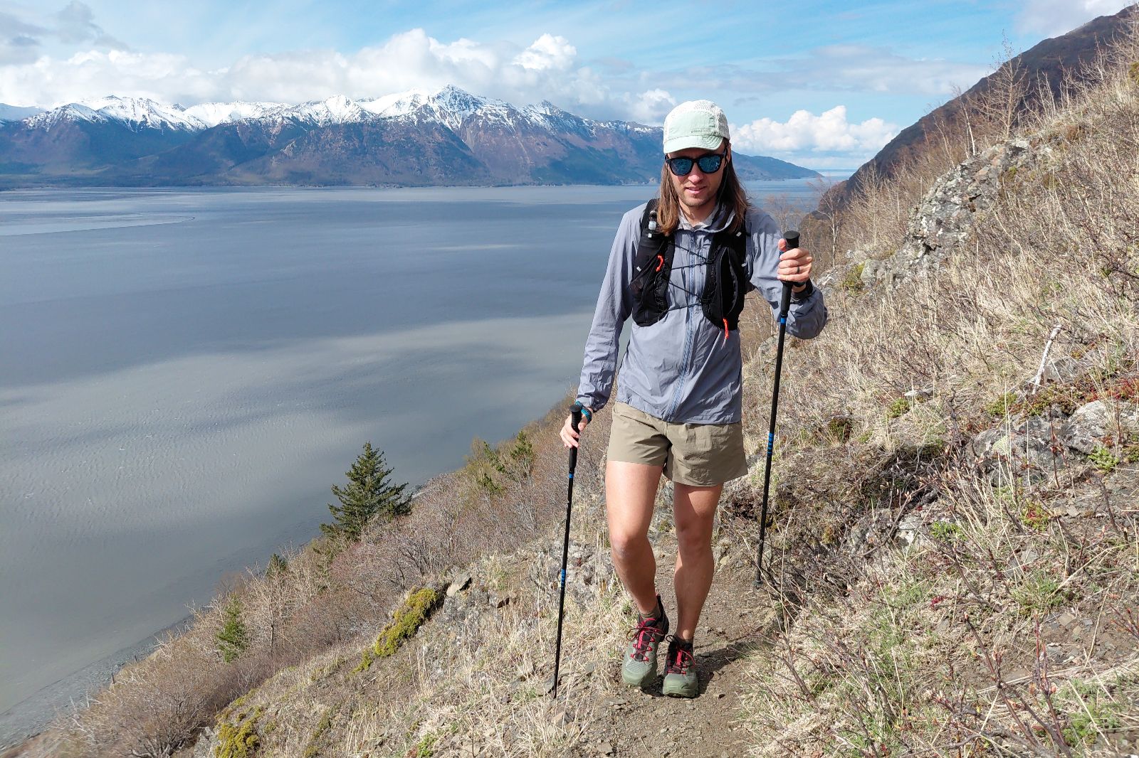 A hiker on a trail above a body of water, with snow-capped mountains in the background.