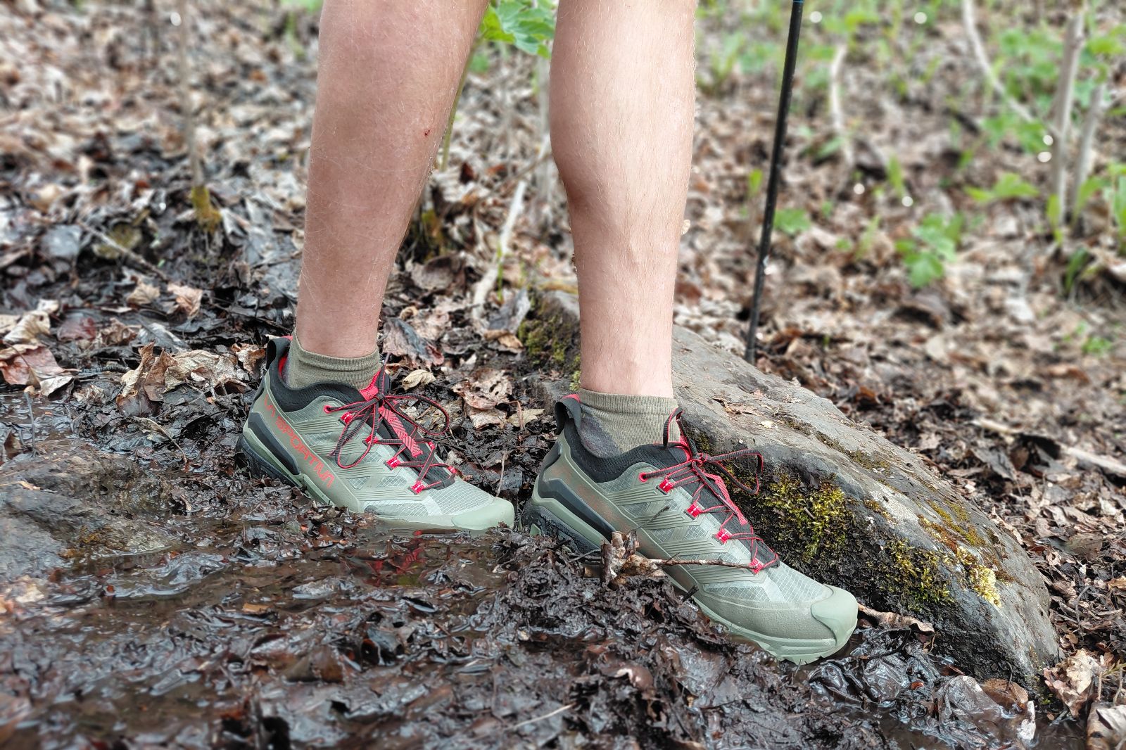 Person wearing olive-green trail shoes with red laces standing on a muddy path.