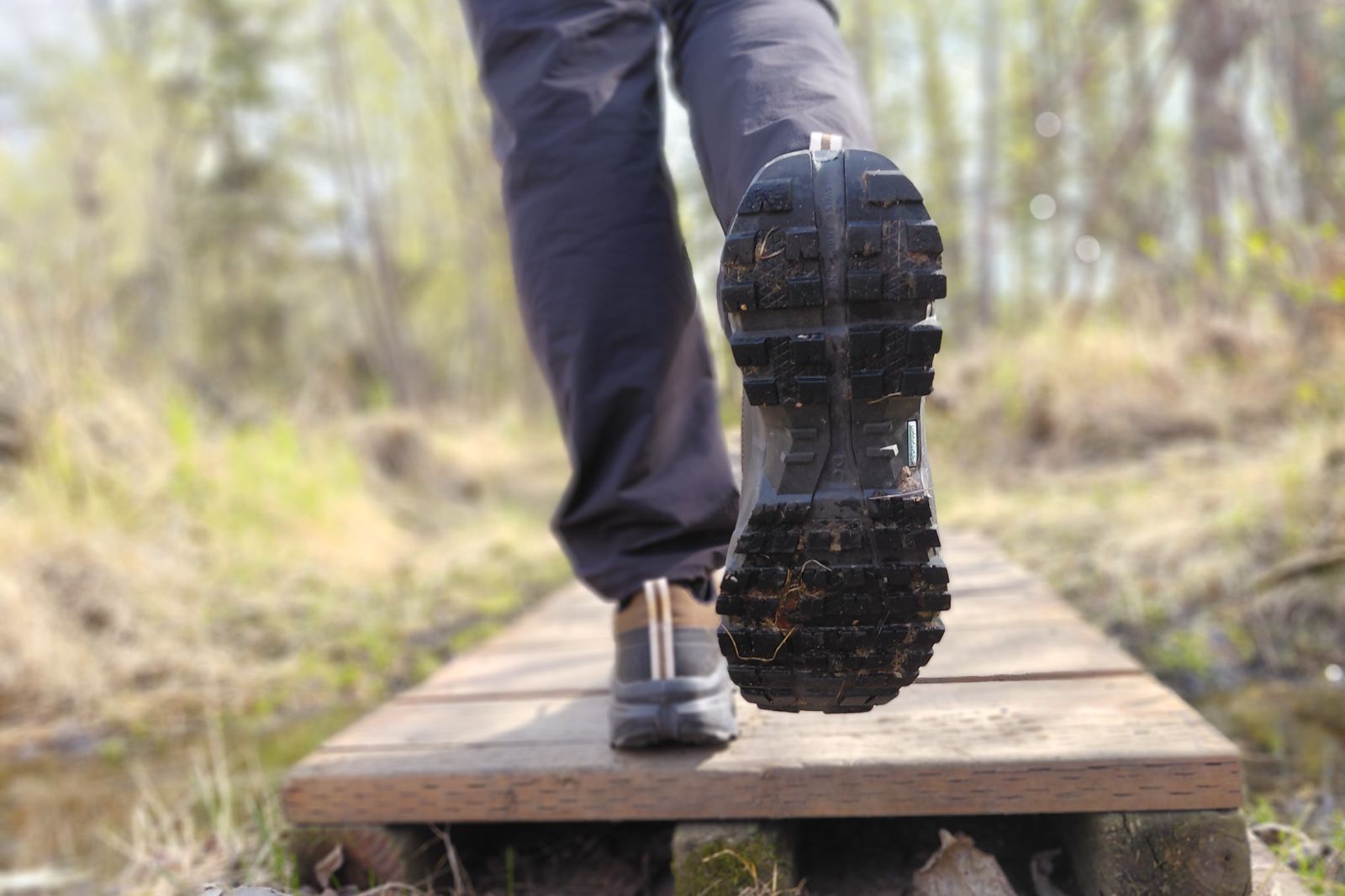 Close-up of a person's shoe with a rugged sole walking on a wooden boardwalk in a natural setting.