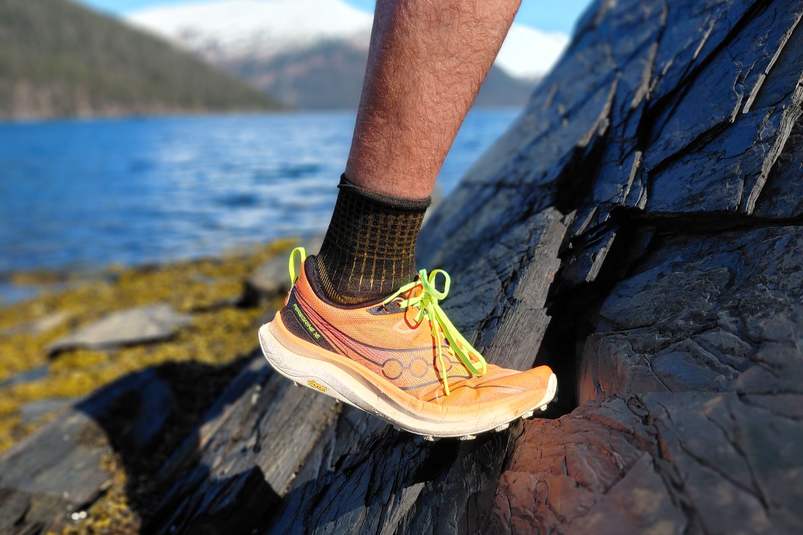 A bright orange running shoe with neon green laces on dark gray rocks by the ocean and mountains.