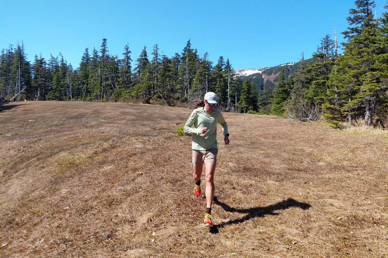 Person running in a meadow with evergreen trees and a snow-capped mountain in the background.
