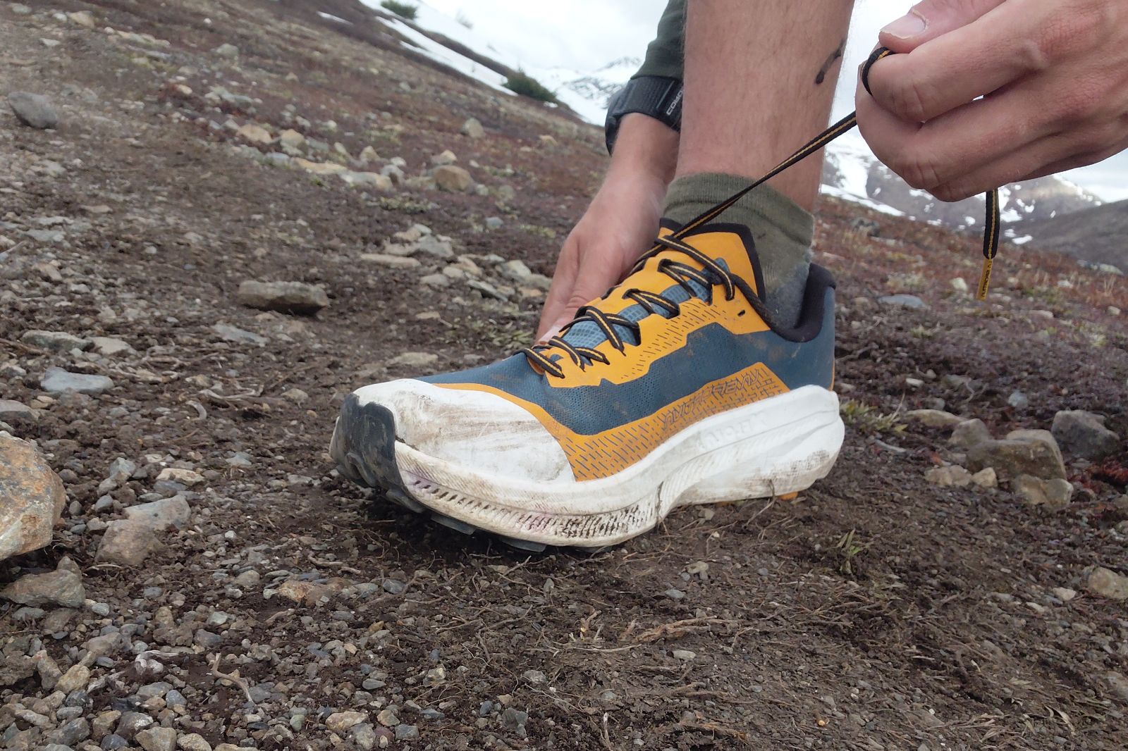 Close-up of someone tying the laces of an orange and blue trail running shoe on rocky ground.