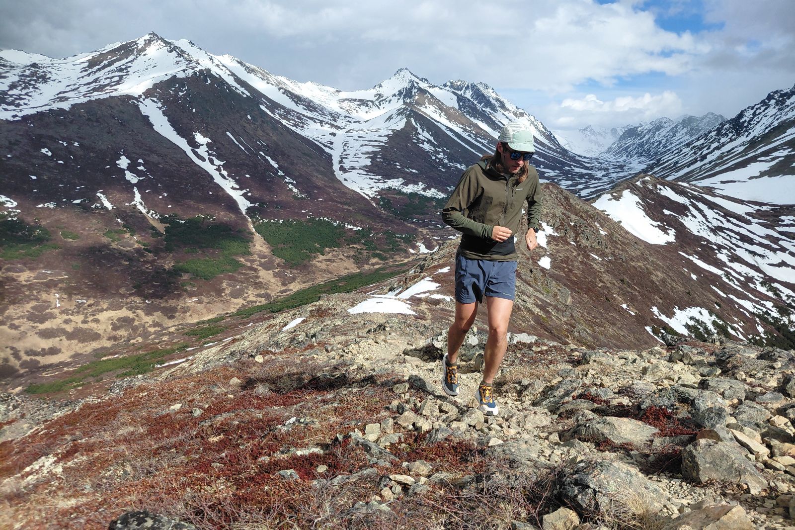 A man runs on a rocky mountain trail with snowy peaks in the background.