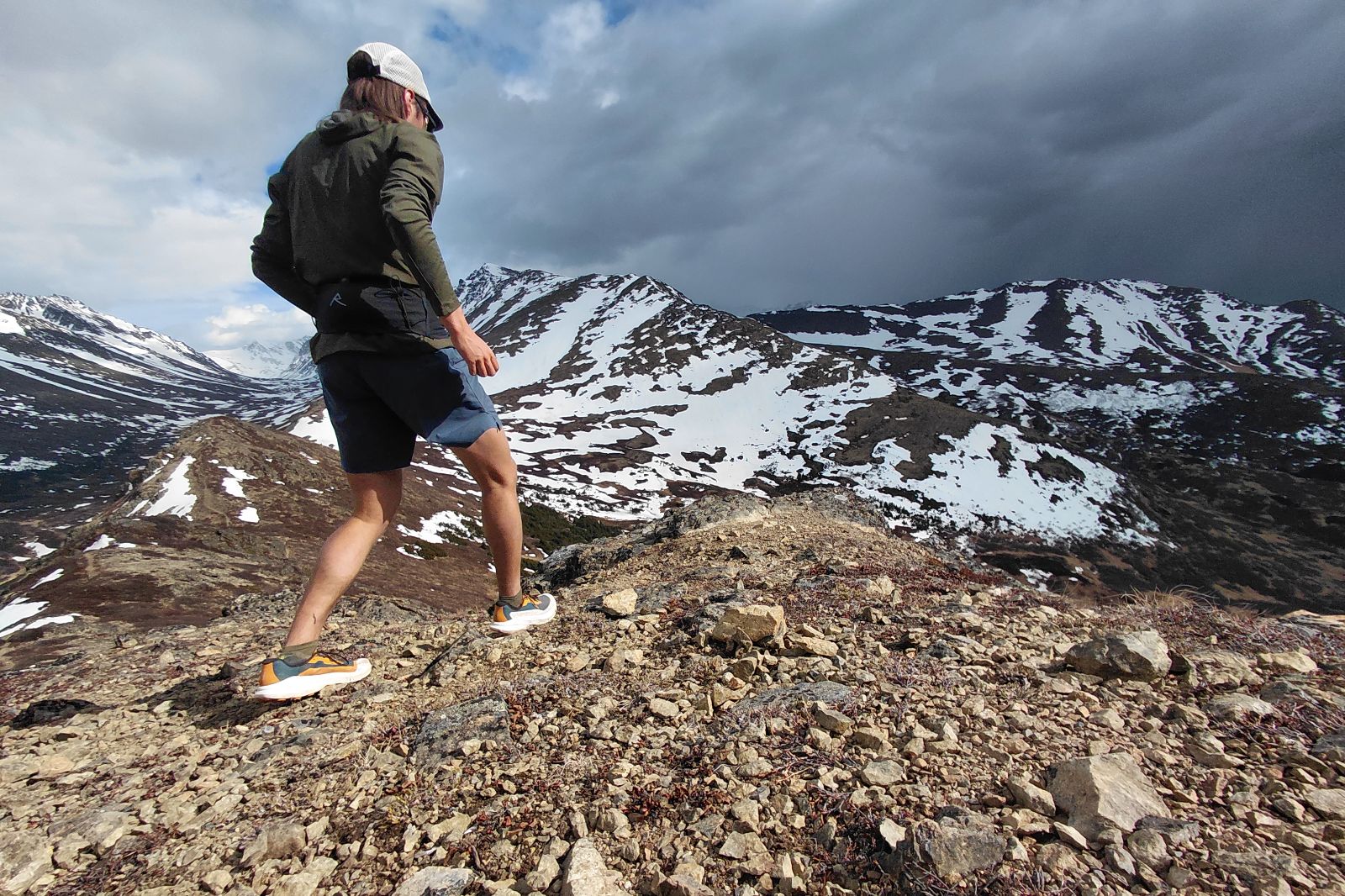A person hikes on rocky terrain with snow-capped mountains in the background.