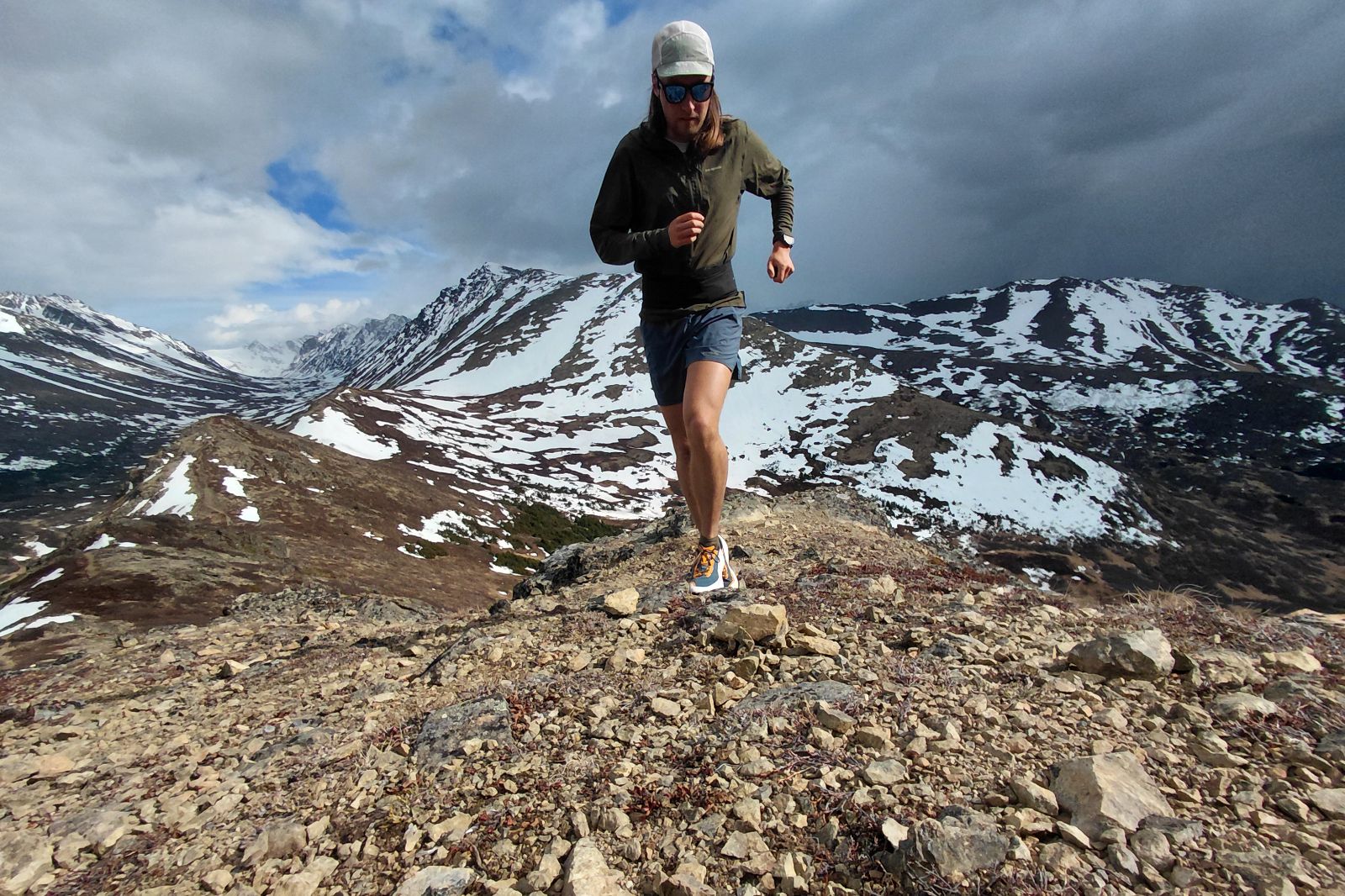 Person running on a rocky mountain trail with snow-capped mountains in the background.