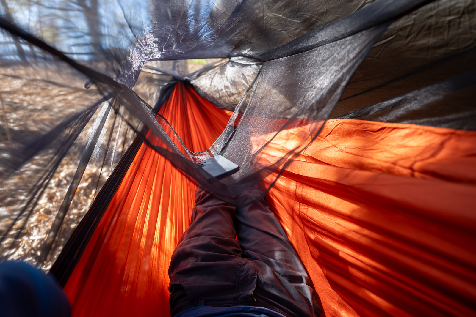 A phone hangs from a mesh pocket in the ceiling of a hammock.