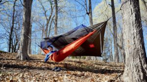 A man hangs in an orange hammock with a tarp behind him in a forest.