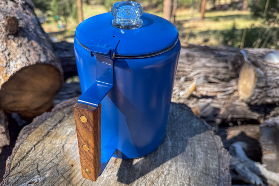 A coffee percolator closed and resting on a tree stump, showing the blue body, wood handle, and glass top knob.