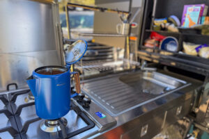 A coffee percolator brewing on a camp stove in an outdoor trailer kitchen setup.