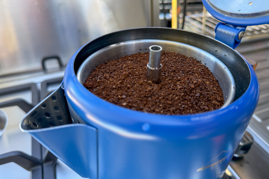 Close-up image of a coffee percolator basket loaded with coffee grounds inside the open blue pot.