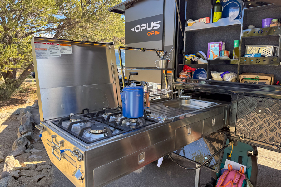 Wide view of a coffee percolator sitting on a two-burner camp stove in an outdoor trailer kitchen.