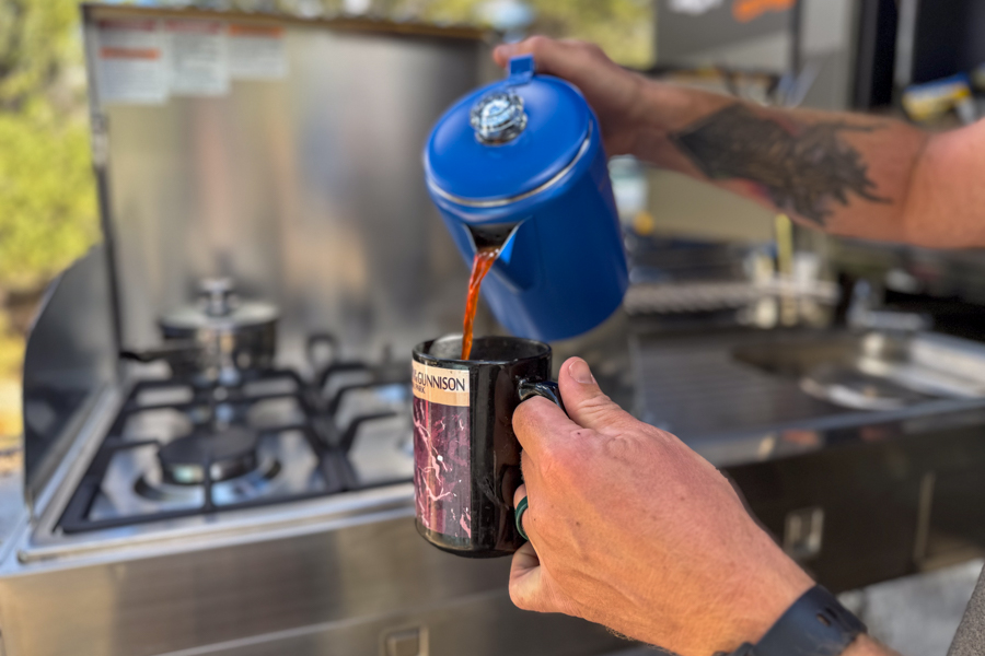 Coffee being poured from a coffee percolator into a camp mug in an outdoor kitchen.