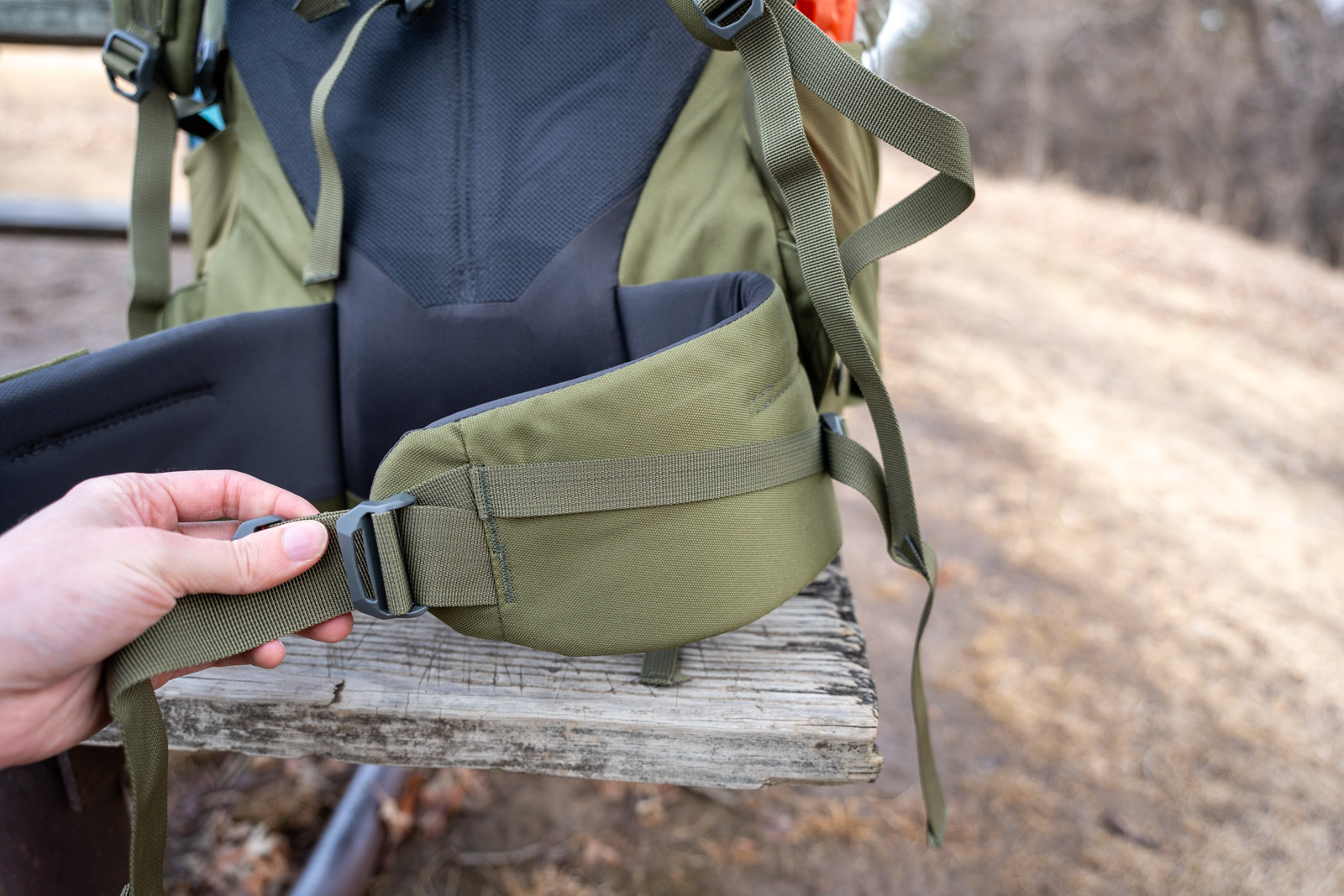A hand holds a green backpacking hip belt connected to a backpack on a picnic table