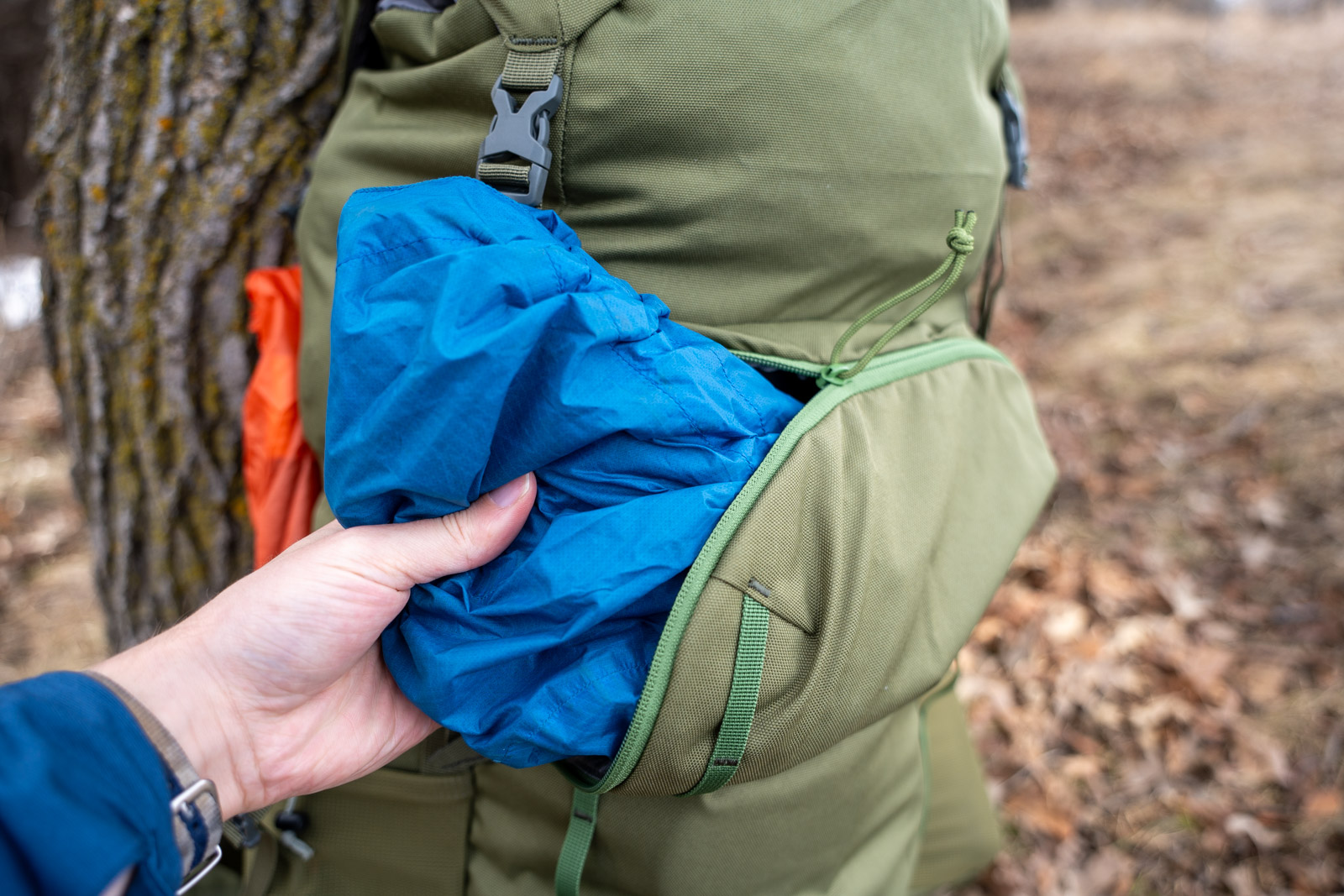A hand pulls a blue jacket out of a zippered pocket on a green backpack