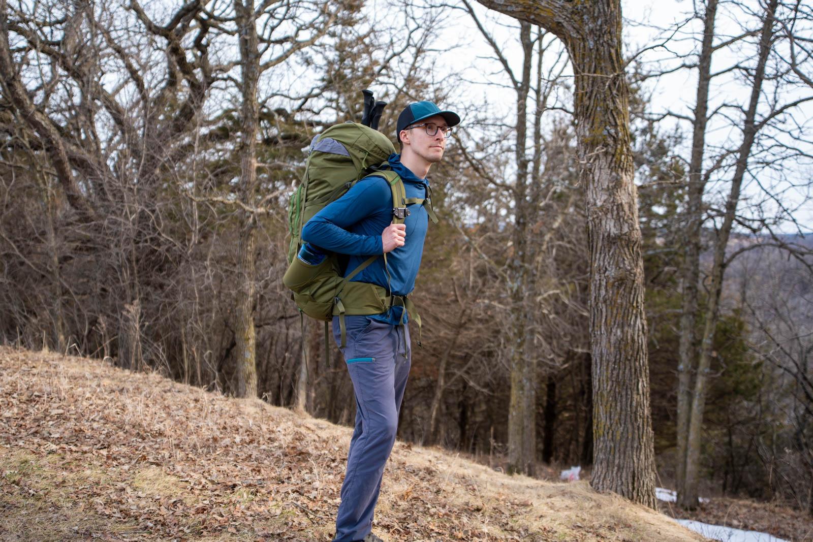 A man wearing blue wears a green backpack in front of a spring forest