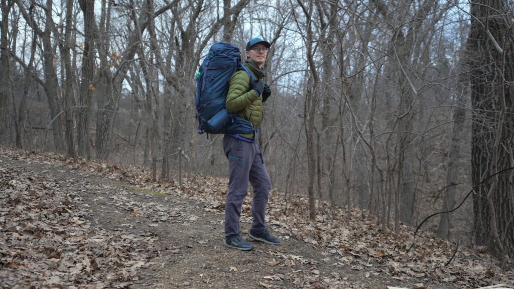 A man in a green jacket wears a blue backpack in the forest