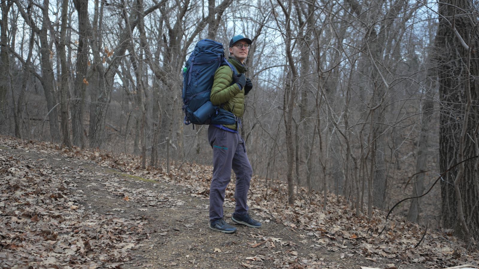 A man in a green jacket wears a blue backpack in the forest