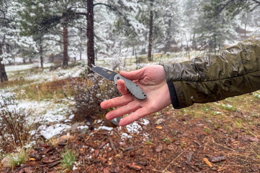 Coast Contra knife held open in one hand during falling snow at a wet campsite.