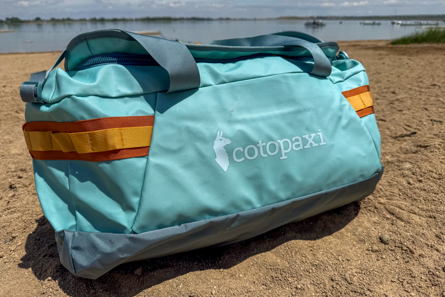 Cotopaxi Allpa Getaway 55L duffel sitting on sand near a lake with tote handles, side stash pocket, and daisy chains visible.