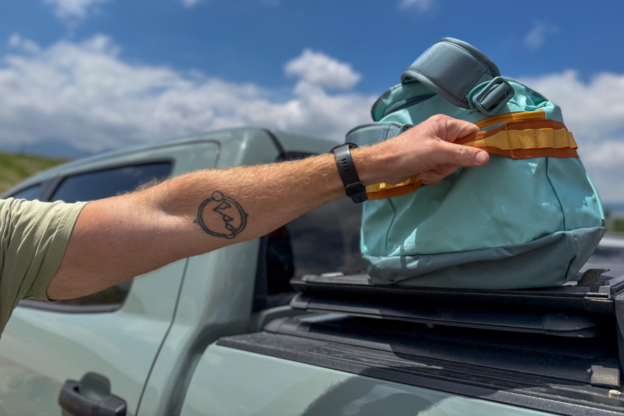 Close-up of a person holding the orange and yellow daisy chain webbing on the Cotopaxi Allpa Getaway 55L near a vehicle.