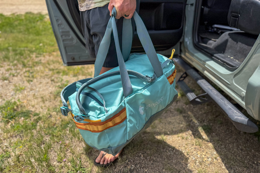 Person carrying the Cotopaxi Allpa Getaway 55L by its tote handles next to an open vehicle door.