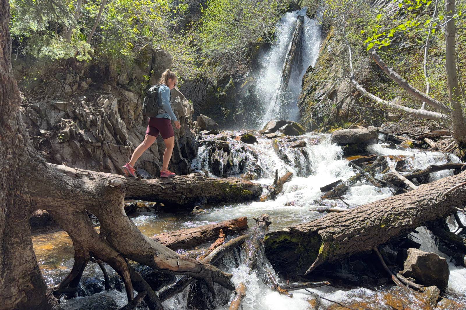 A woman wearing thigh-length hiking shorts crosses a log in front of a waterfall.