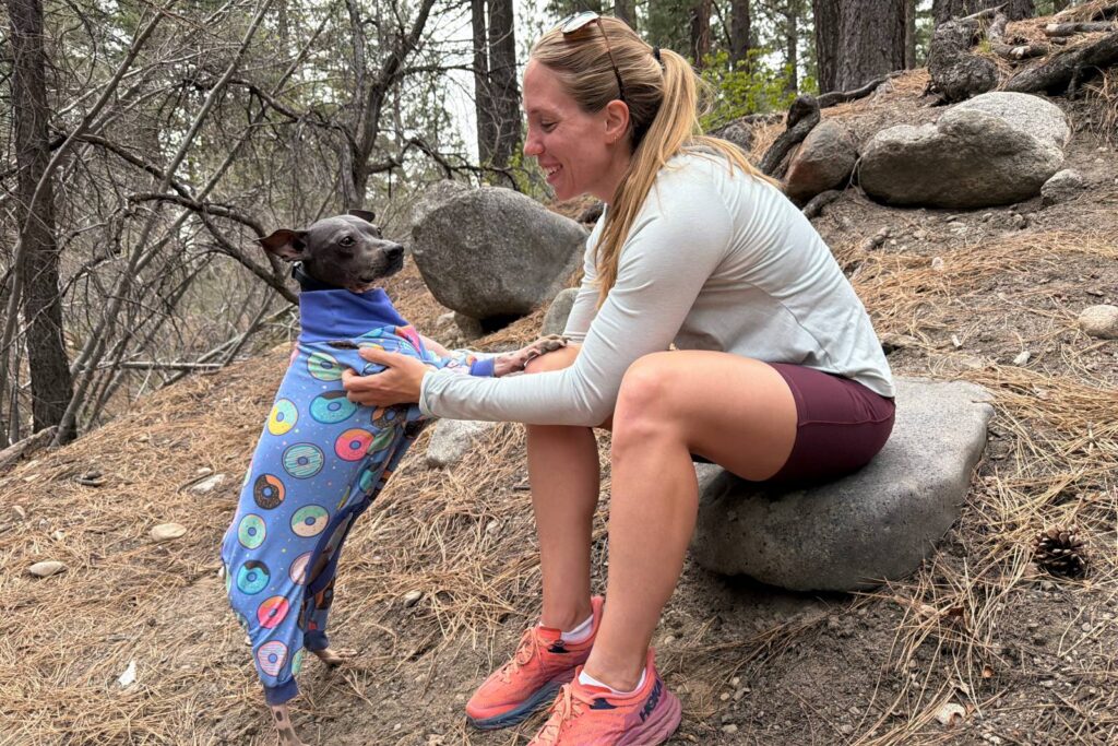 A woman wearing hiking shorts is sitting on a rock and reaching forward to support a dog, who is standing on his hind legs with his paws on her knee.