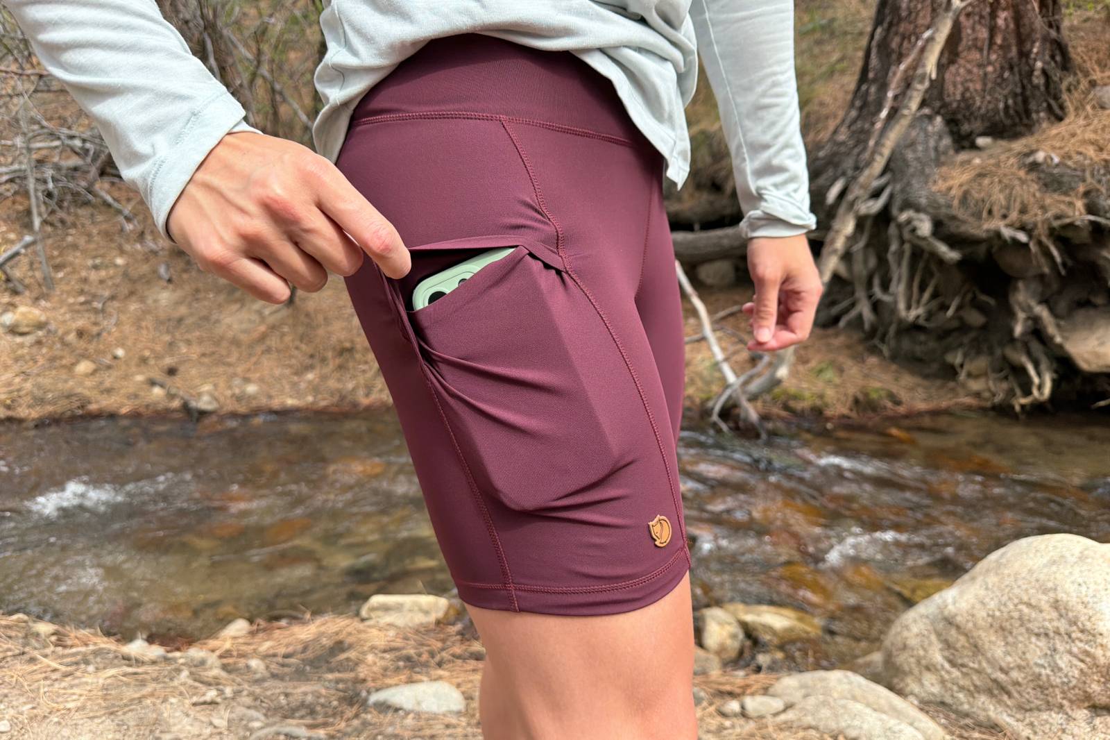 Close-up image of a woman reaching down to reveal a smartphone inside the pocket of her hiking shorts.