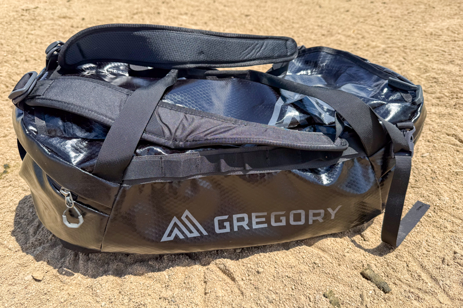Black Gregory Alpaca 60 duffel sitting on sand near a lake with shoulder straps and grab handles visible.