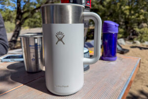 A French press sitting on a camp table with its light gray handle and powder-coated body visible.