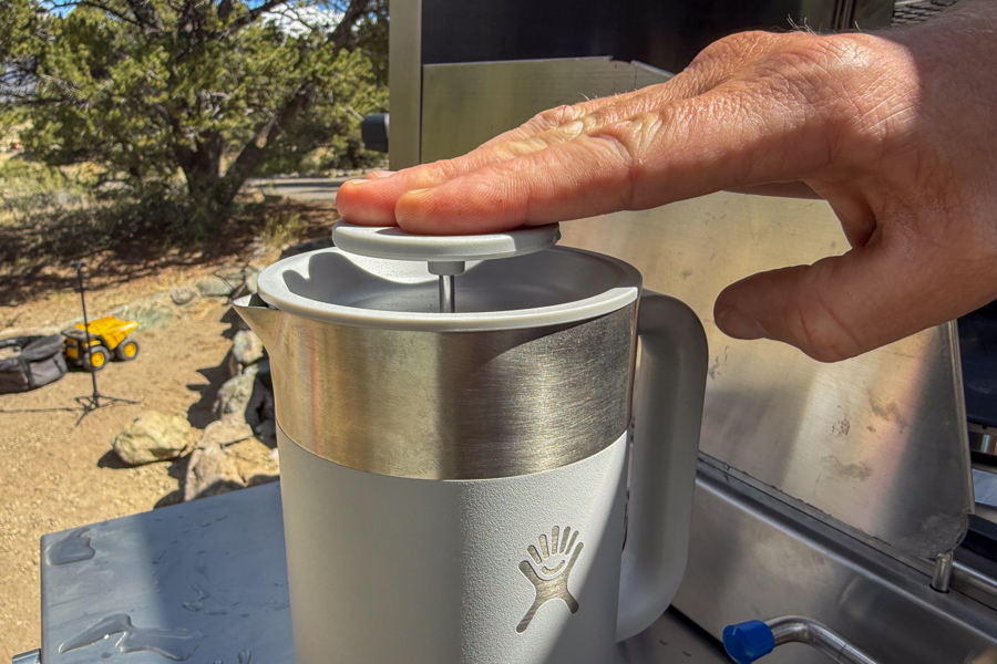 Hand pressing the plunger down on the a French Press lid at a camp kitchen.