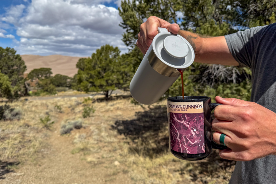Coffee being poured from a French press into a ceramic camp mug with Great Sand Dunes National Park in the background.