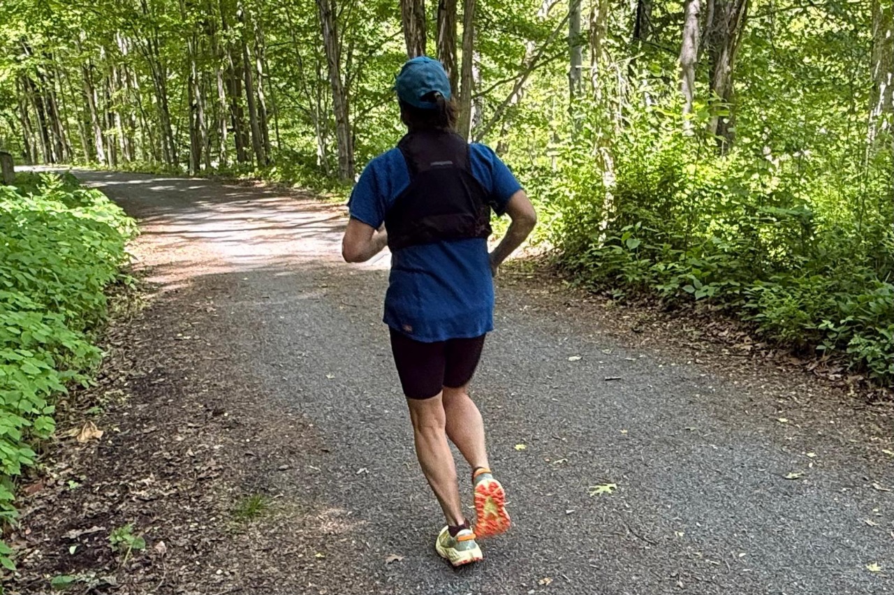 A woman running while wearing a black running vest. She is on a flat, forested trail.