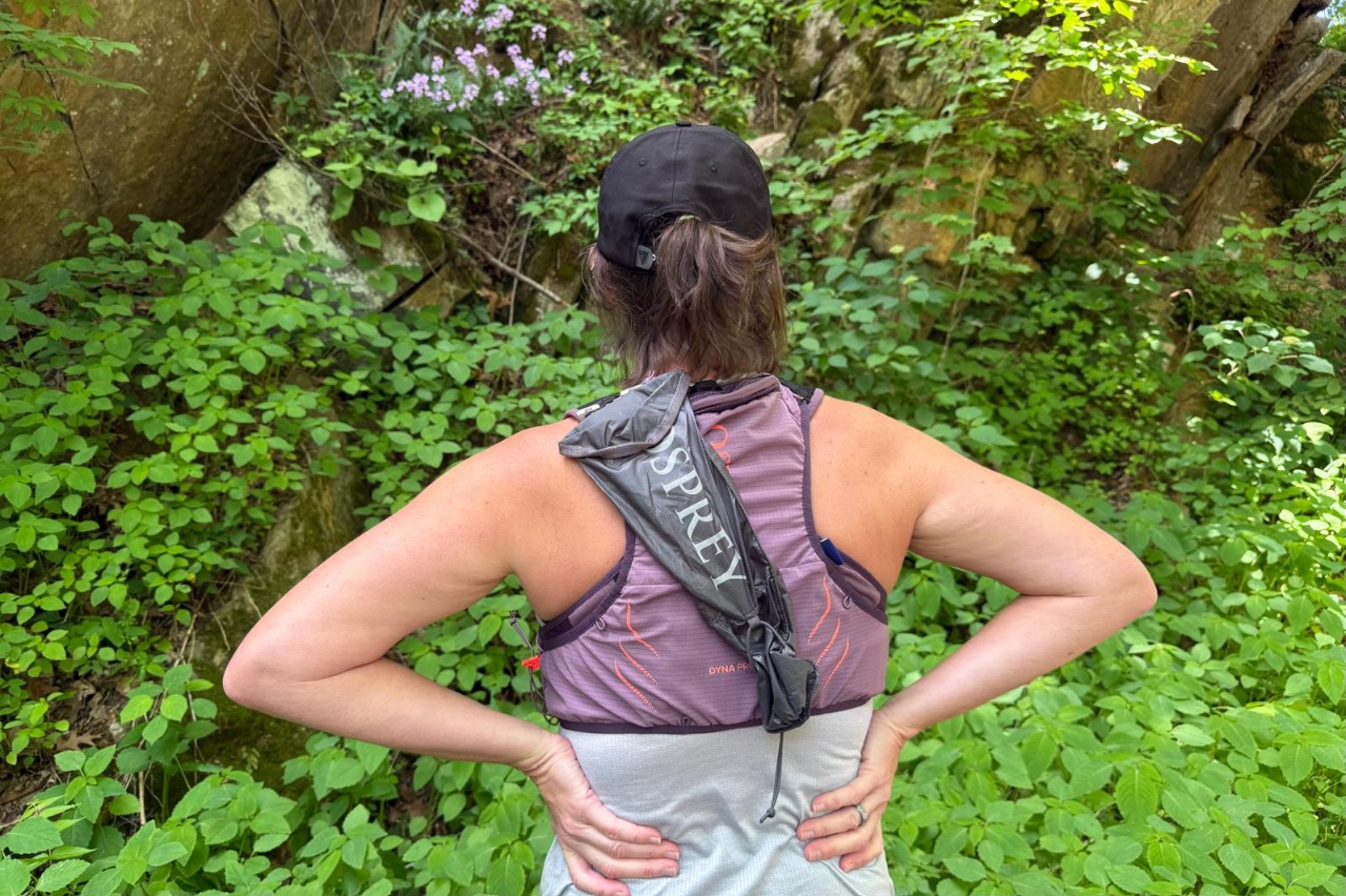 A woman stands with her hands on her hips wearing a purple running vest with a trekking pole quiver attached at the back.