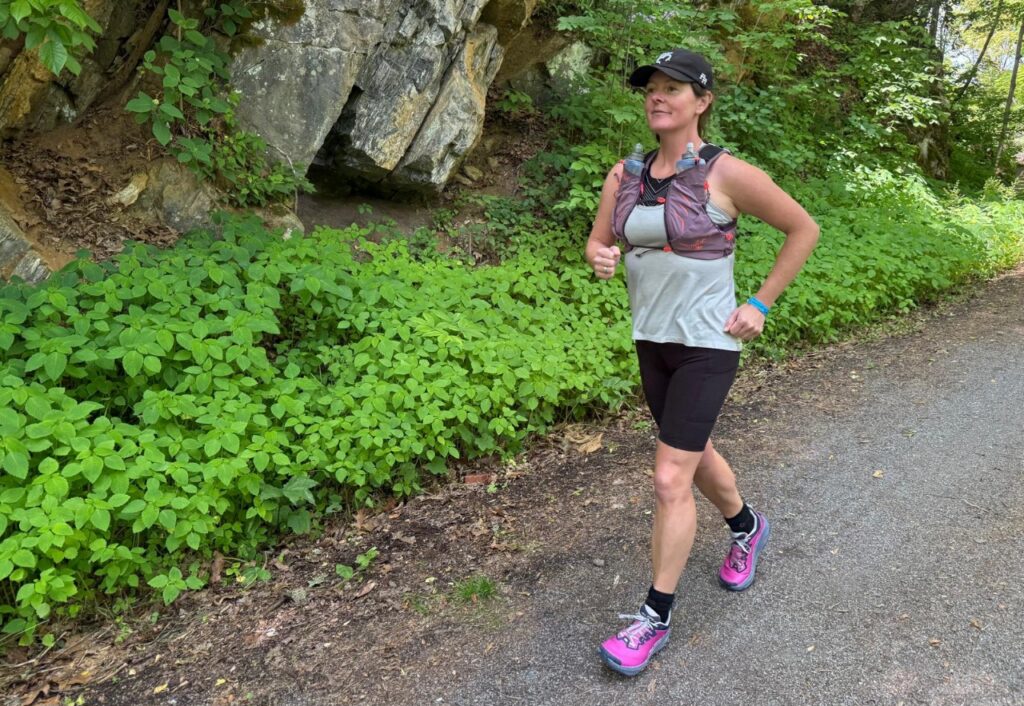 Woman running on a flat, forested trail wearing a purple hydration vest.