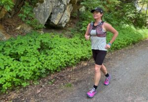 Woman running on a flat, forested trail wearing a purple hydration vest.