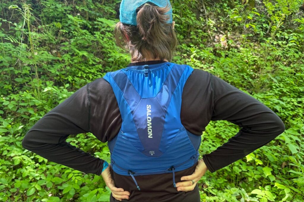 Close-up image of a woman standing in front of greenery wearing a blue running vest. The back of the vest is visible, showcasing the large main compartment.