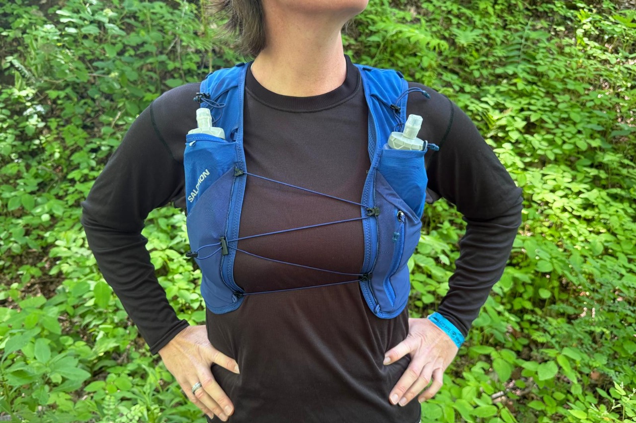 Close-up image of a woman wearing a blue running vest. She is standing in front of greenery with her hands on her hips.