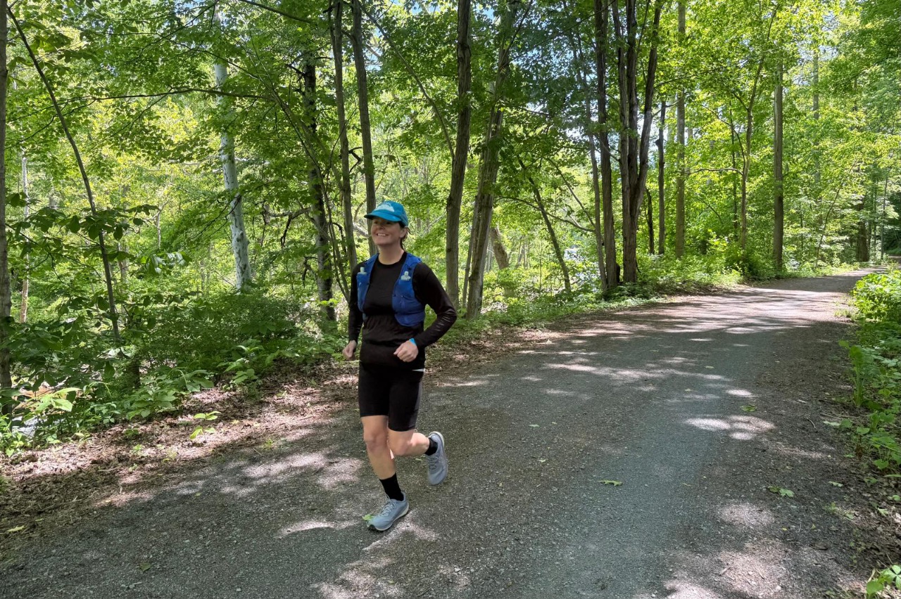 A woman running on a flat, forested trail wearing a blue running vest.