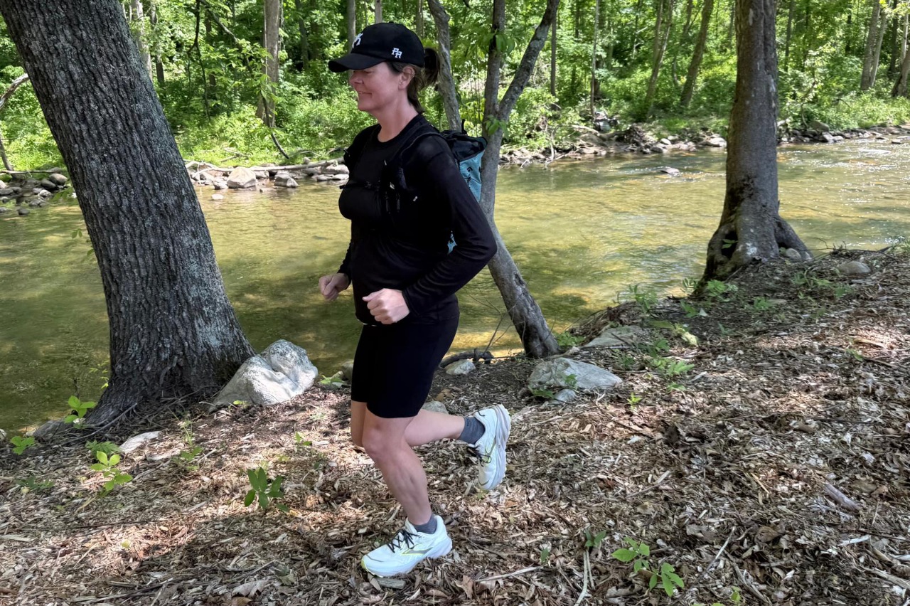 Woman wearing a blue day pack/running vest running along a forested trail.
