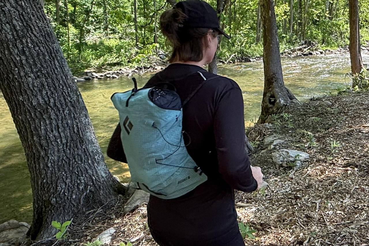 Woman wearing a blue day pack/running vest while running on a forested trail.
