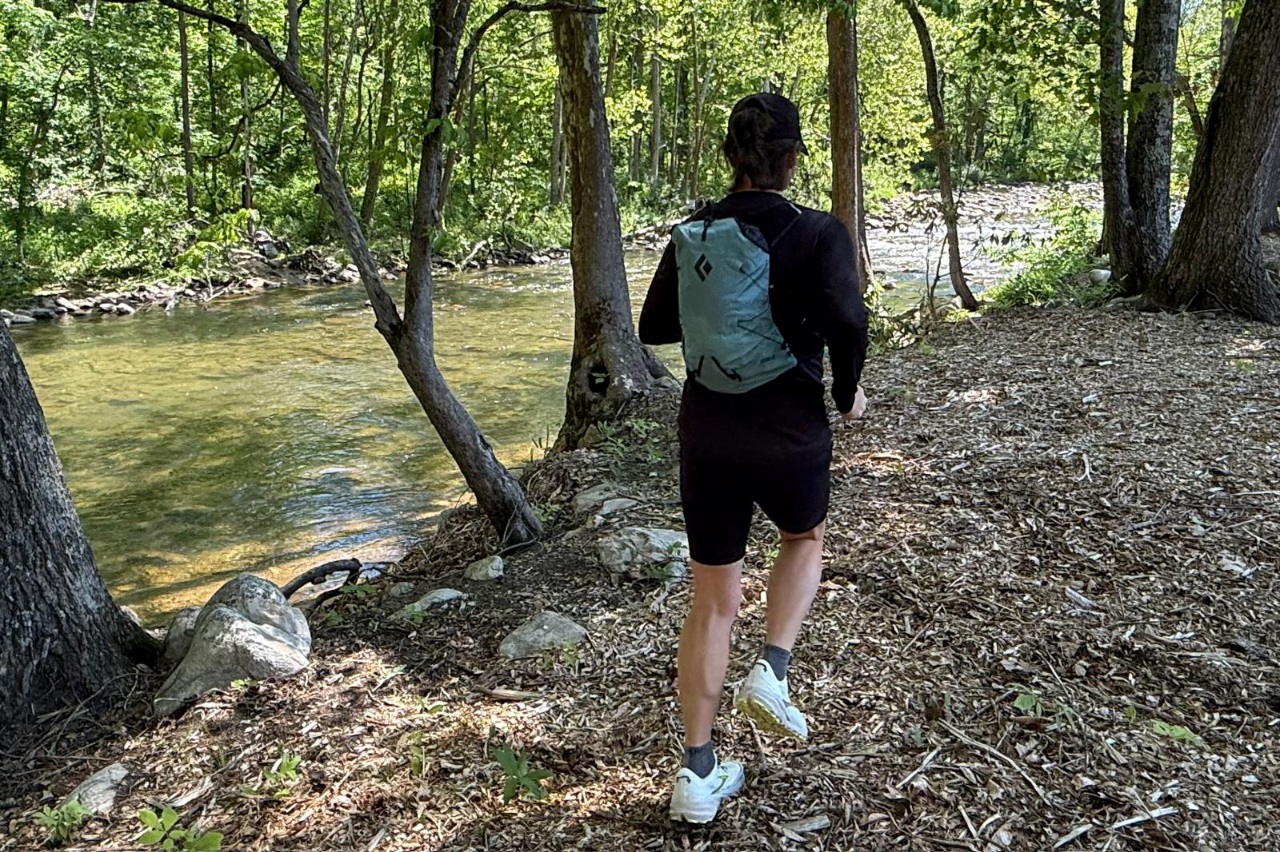 Woman wearing a blue day pack/running vest running along a forested trail with a river on one side.