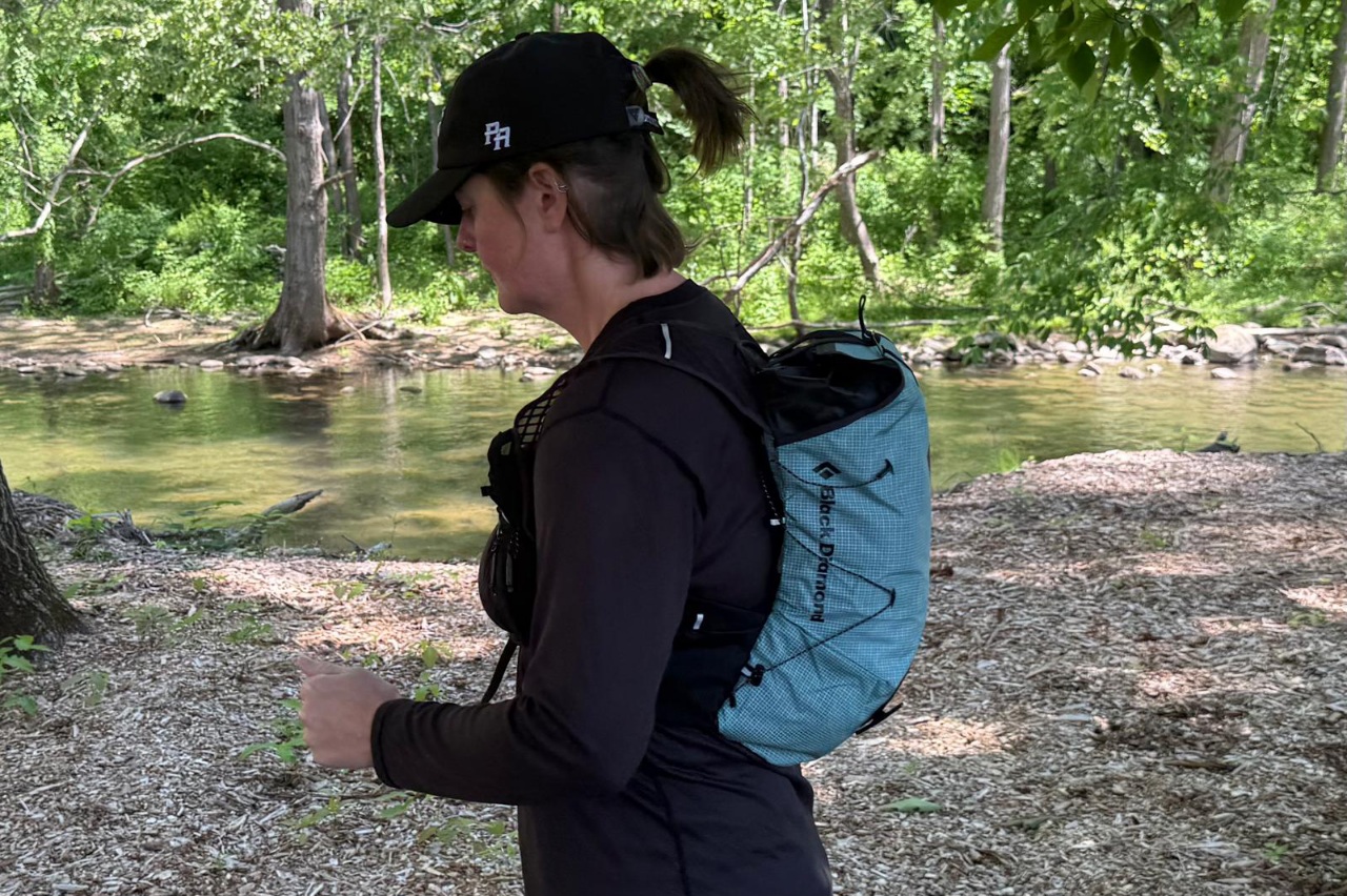 Side view of a woman running with a blue day pack/running vest. There are trees and greenery in the background.