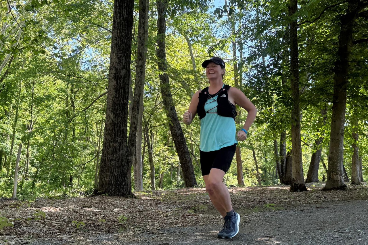 Woman running on a forested trail wearing a black running vest.