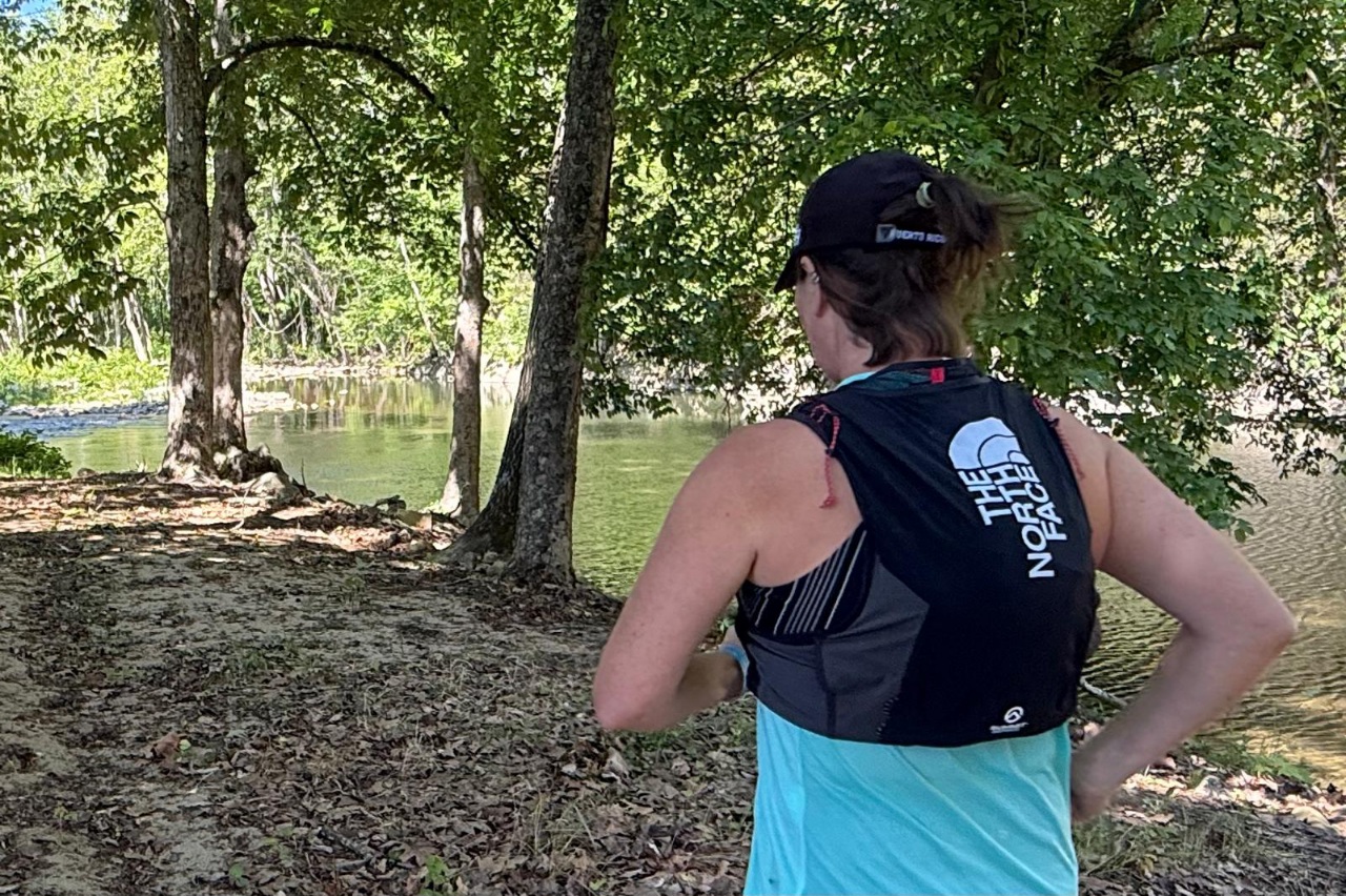 Woman wearing a black running vest running on a flat travel with trees and a river nearby.