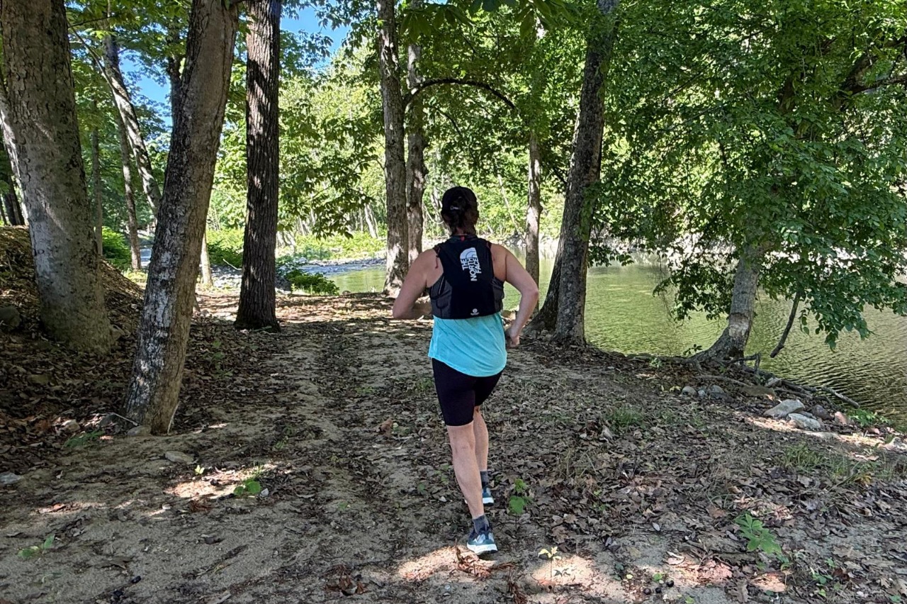 A woman wearing a black running vest runs along a forested trail.